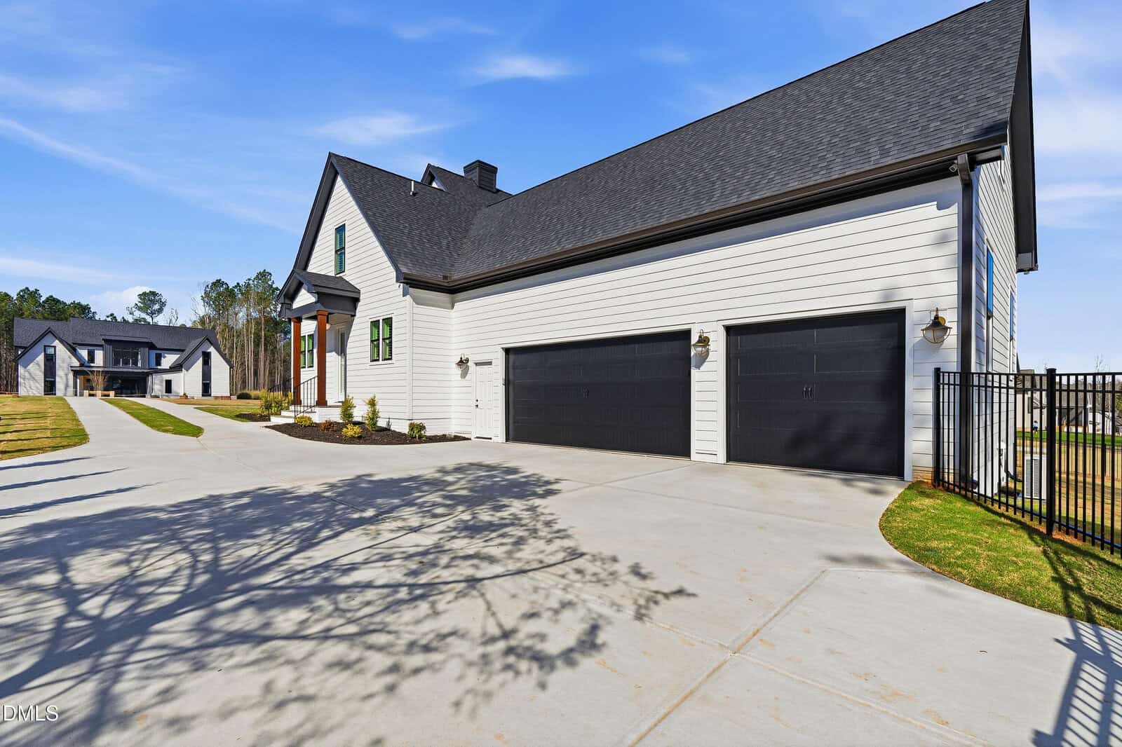 Modern white house with black trim, a three-car garage, large driveway, landscaped yard, and a smaller house visible in the background under a clear blue sky.
