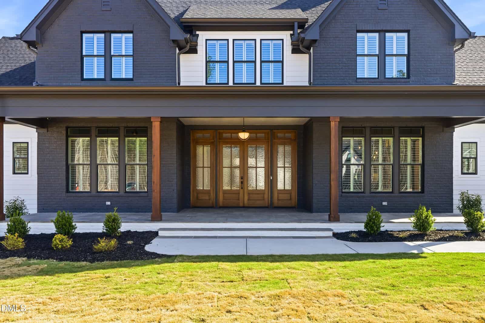 A modern two-story house with dark gray brick, large windows, a covered porch with wooden columns, and wooden double front doors. Small bushes line the front walk, and the lawn is neatly manicured.
