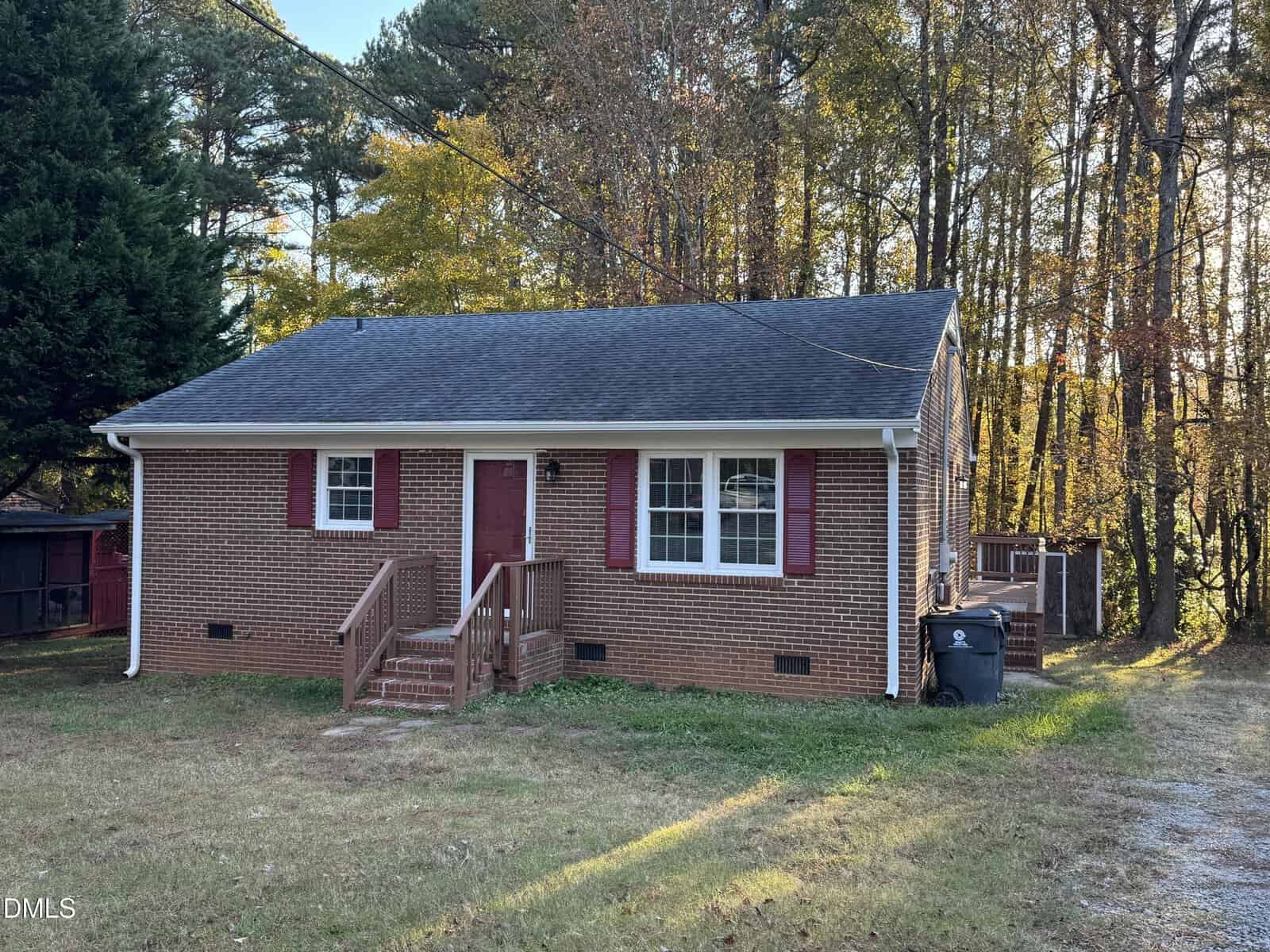 A small brick house with red shutters and door stands at 818 Wakefield Avenue in Henderson, featuring a simple front porch and steps leading to a grassy yard. Tall trees surround the house, casting light shadows on the scene.