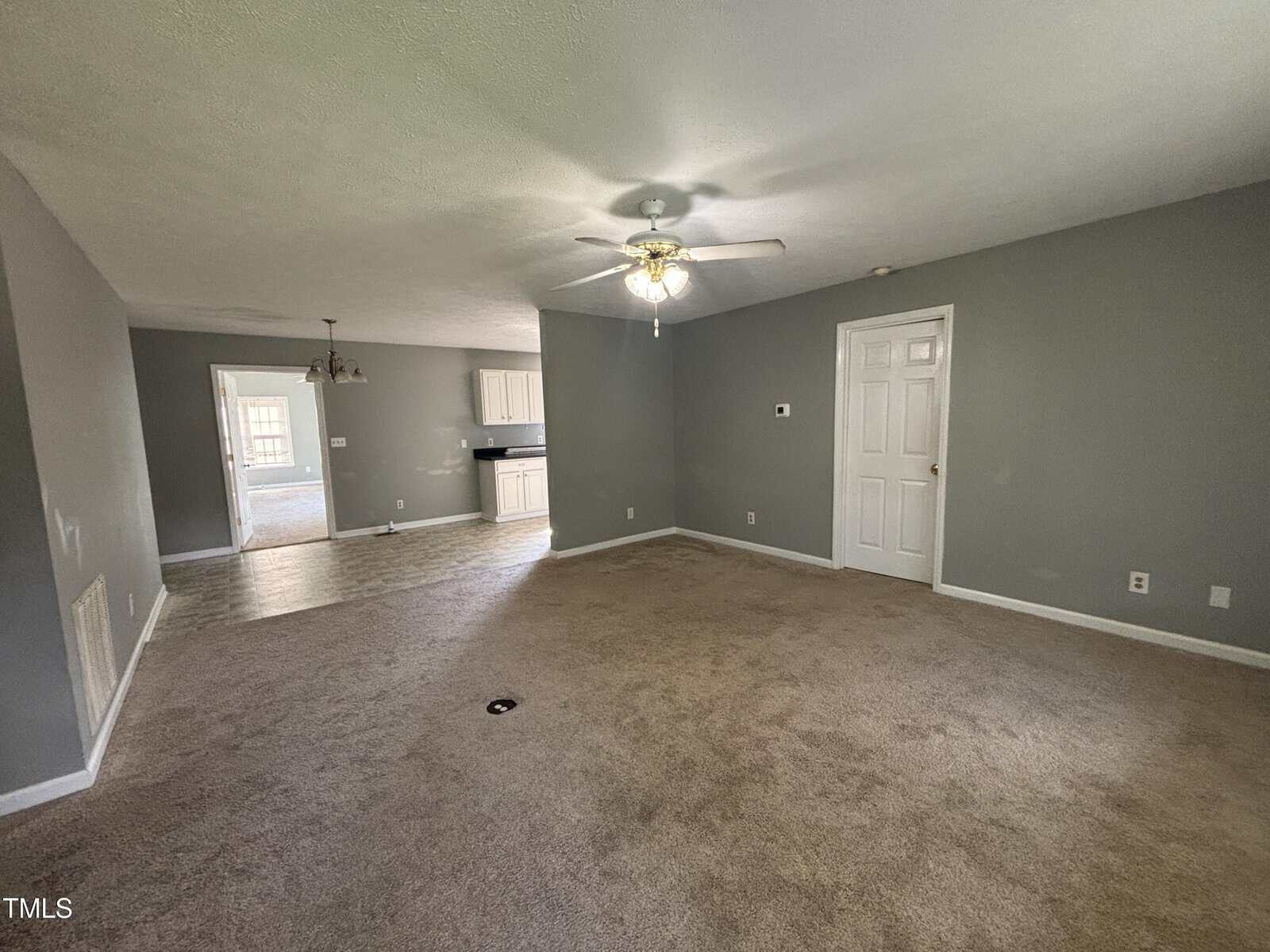 A spacious living room with beige carpet, light gray walls, and a ceiling fan. The room opens to a tiled dining area and a kitchen with white cabinets. Multiple doors are visible along the walls.