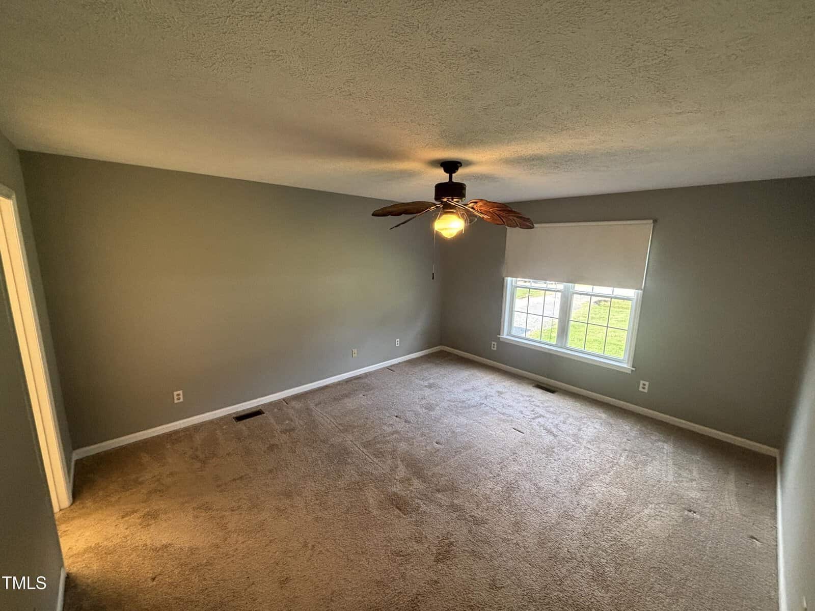 Empty carpeted room with gray walls, a ceiling fan with leaf-shaped blades, a large window with a white blind, and natural light coming in. Electrical outlets and a vent are visible along the walls.