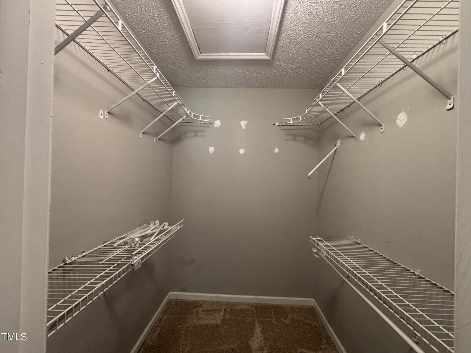Empty walk-in closet with wire shelving on three walls, patched holes visible on the back wall, a ceiling hatch, and carpeted floor. The walls are painted a light gray color.