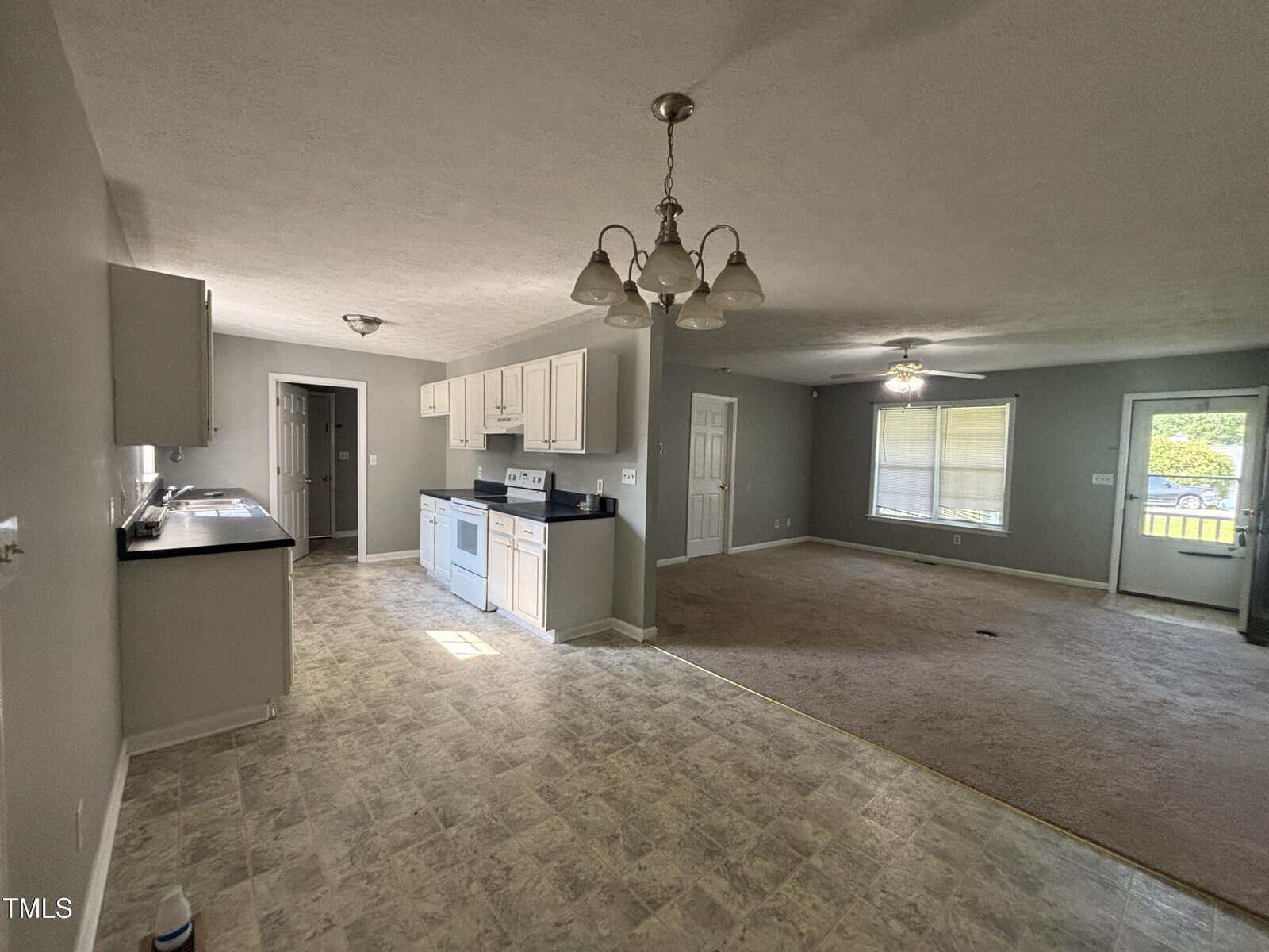 Open-plan kitchen and living room with light gray walls, white cabinets, and blue appliances. The kitchen has tile flooring, while the living area is carpeted. Large windows let in natural light.