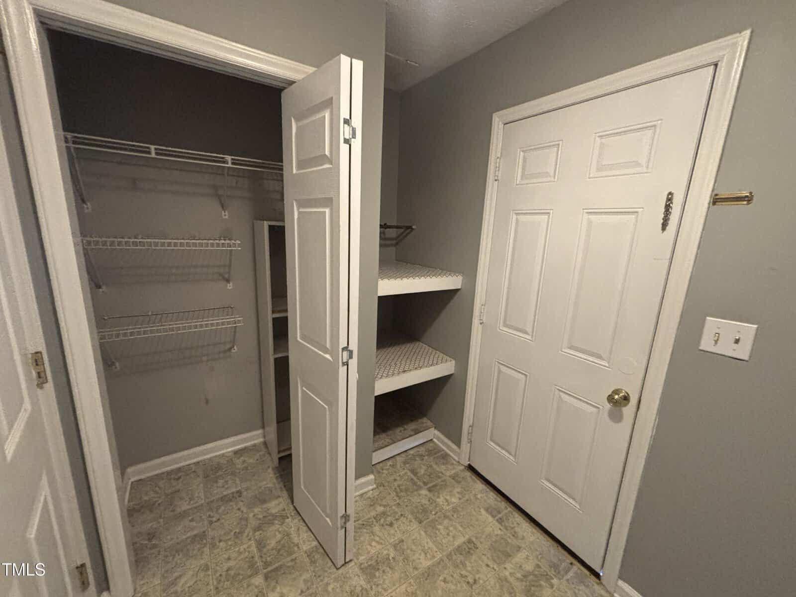 A small room with gray walls, two white doors, wire shelving on the left, and corner shelves on the right. The floor has a gray patterned tile.