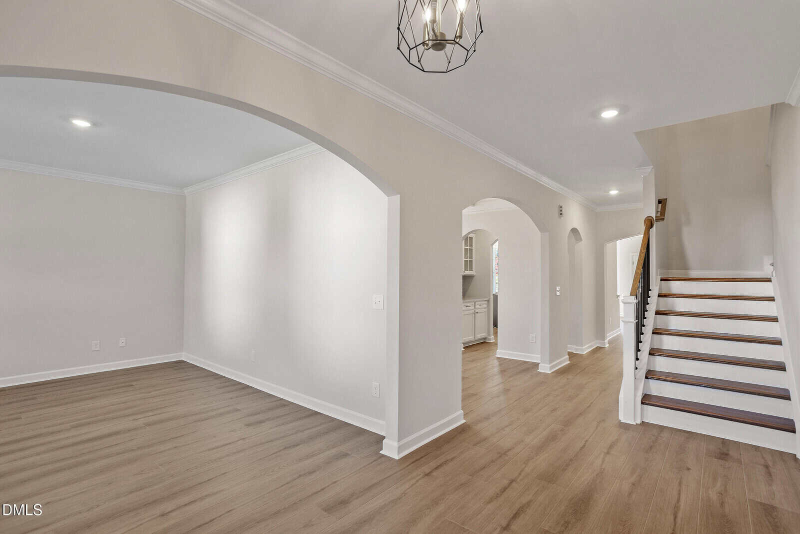 A view of a modern home interior with light-colored walls, wood flooring, white trim, arched entryways, a staircase with a dark handrail, and a geometric ceiling light fixture.