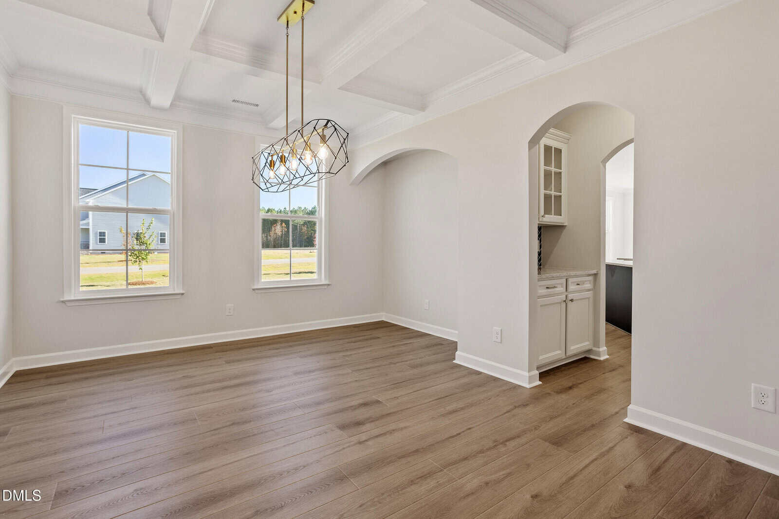 A bright, empty dining room with light-colored walls, large windows, wood flooring, a modern geometric chandelier, coffered ceiling, and an arched doorway leading to a built-in cabinet area.