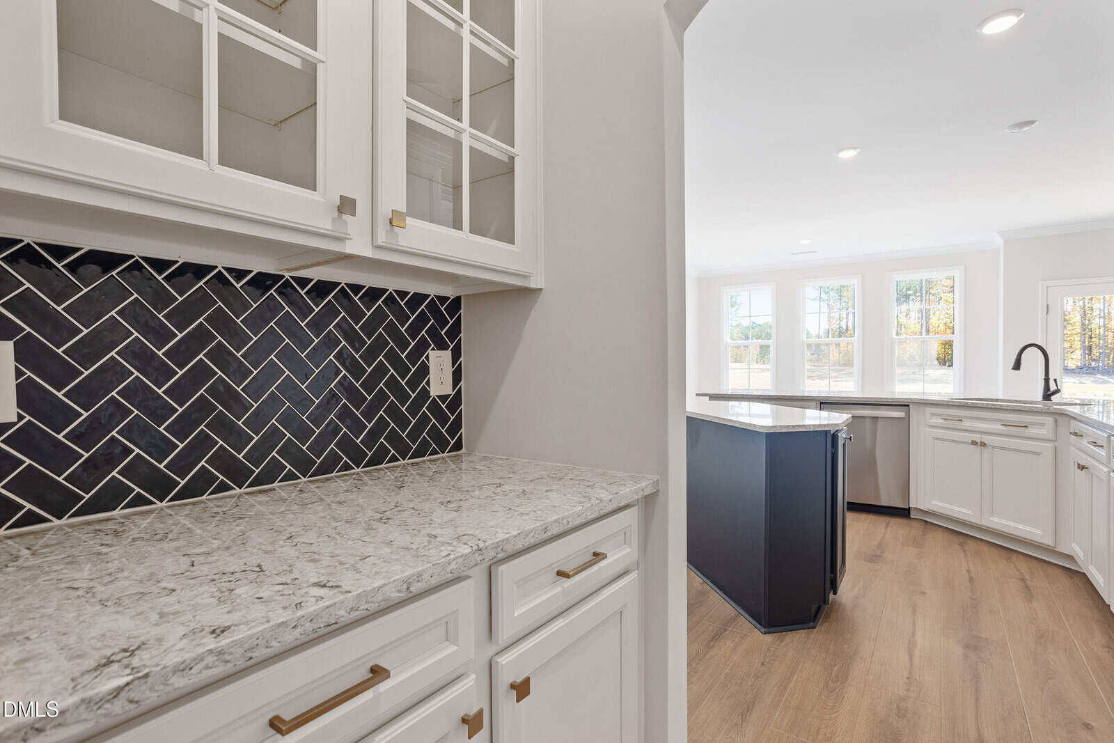 A modern kitchen features white cabinets with brass handles, a marble countertop, a black herringbone tile backsplash, light wood flooring, and an island with a black base. Large windows allow natural light to fill the space.