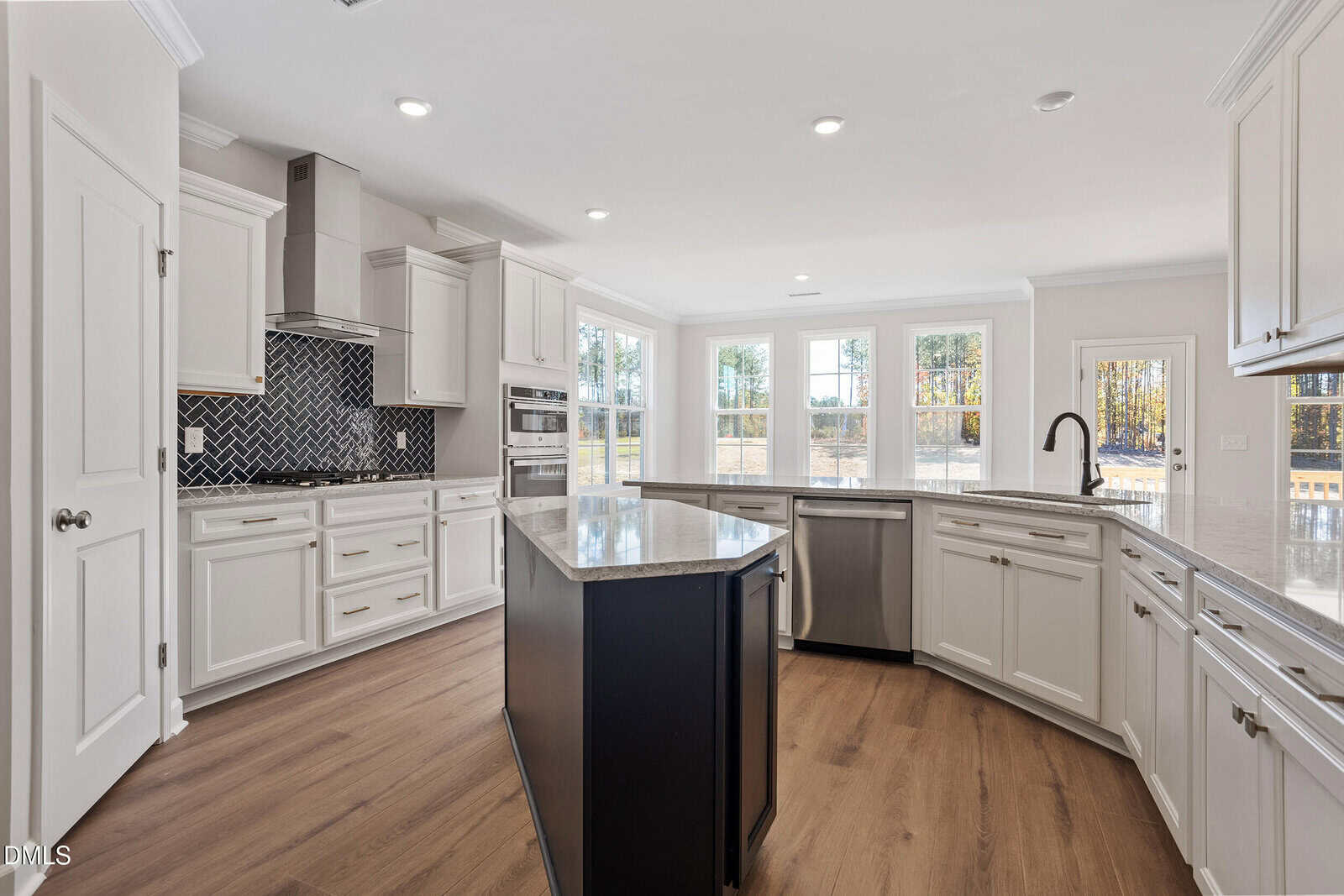 A modern kitchen with white cabinets, stainless steel appliances, a black island with a marble countertop, a herringbone tile backsplash, wood flooring, and large windows letting in natural light.