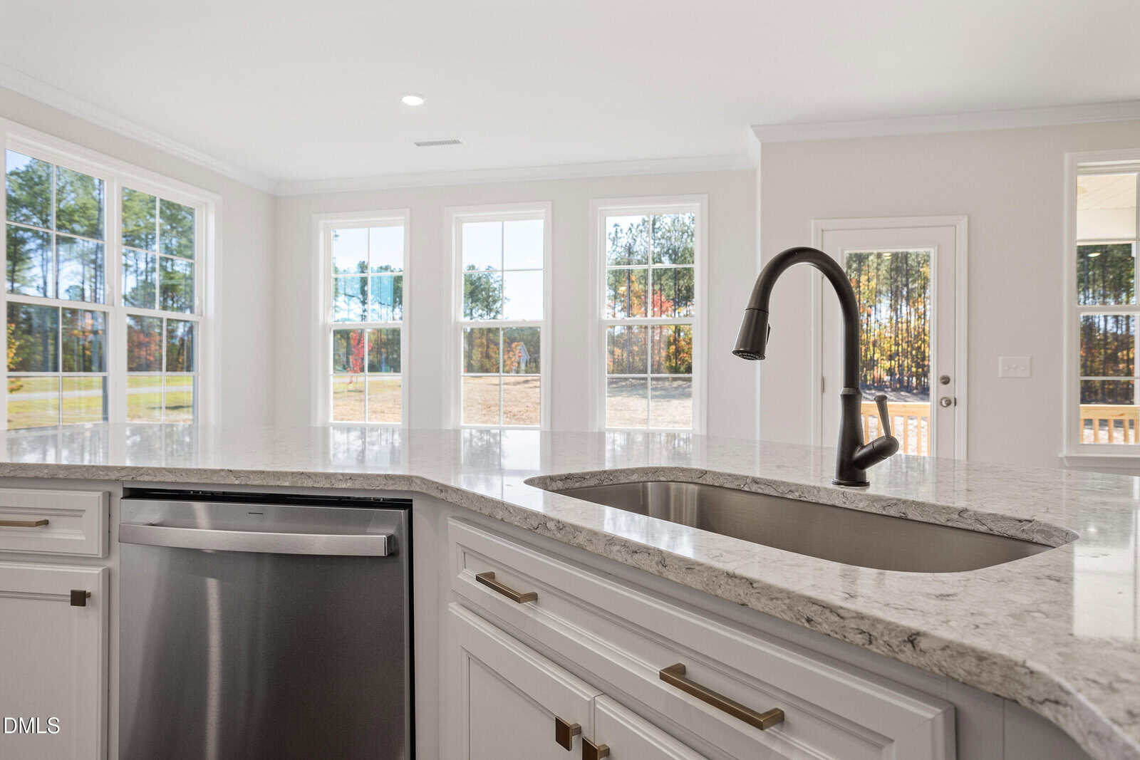 A modern kitchen with white cabinets, a stainless steel dishwasher, a marble countertop, and a built-in sink with a black faucet. Large windows and a glass door let in natural light and offer a view of trees outside.