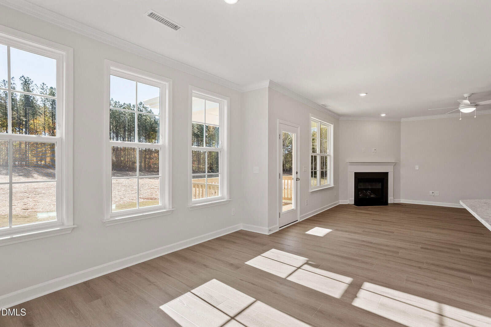 A bright living room with large windows, light-colored walls, and wood flooring. The room features a fireplace, ceiling fan, and a glass door leading outside. Sunlight streams in, casting shadows on the floor.