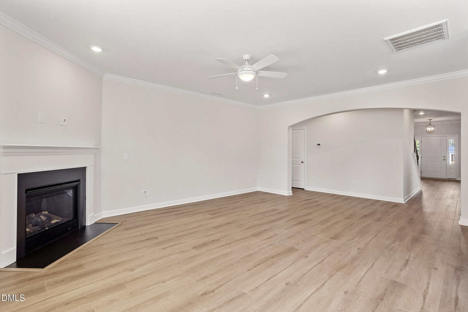 A spacious, empty living room with light wood flooring, a corner fireplace, a ceiling fan with a light, white walls, and an arched doorway leading to a hallway and a front door.