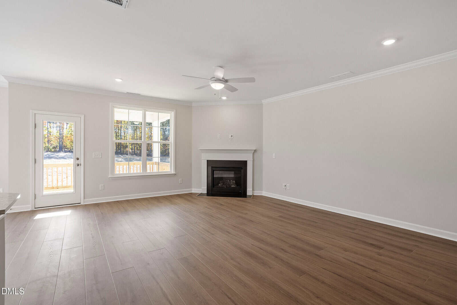 Empty living room with light-colored walls, wood flooring, a ceiling fan, a fireplace, a large window, and a door leading outside. Natural light enters through the window and door, creating a bright, open space.