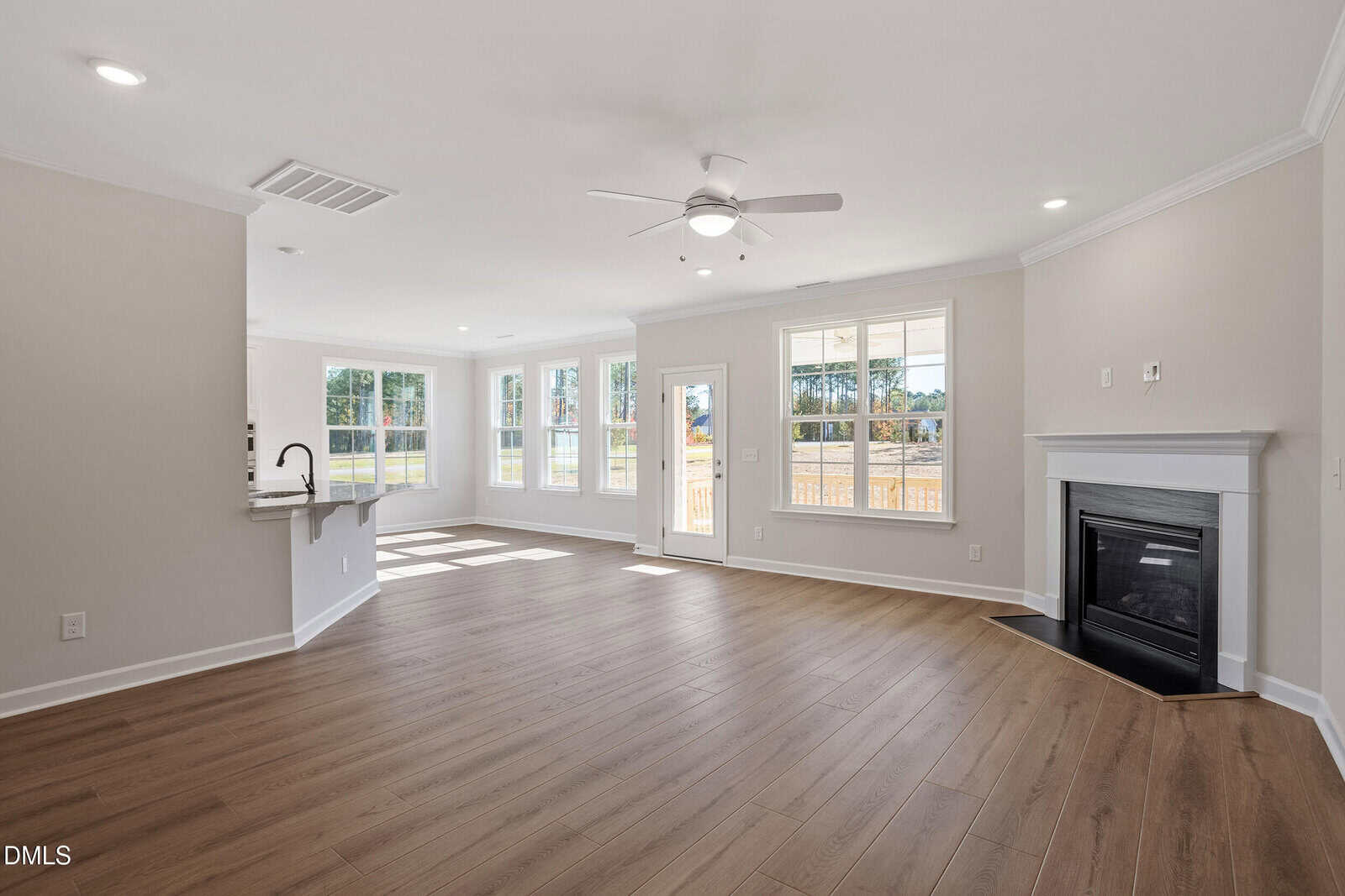 Bright, empty living room with light wood flooring, large windows, a ceiling fan, a fireplace, and an open layout leading to a kitchen with a breakfast bar. Door leads outside. Walls are painted light neutral colors.