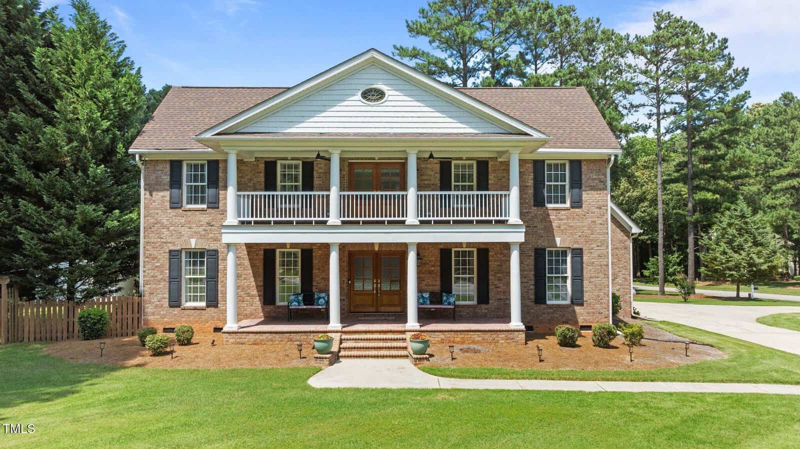Two-story brick house with white columns, a front porch, and an upper balcony. It has large wooden double doors, symmetrically placed windows, and is surrounded by green grass, trees, and a clear blue sky.