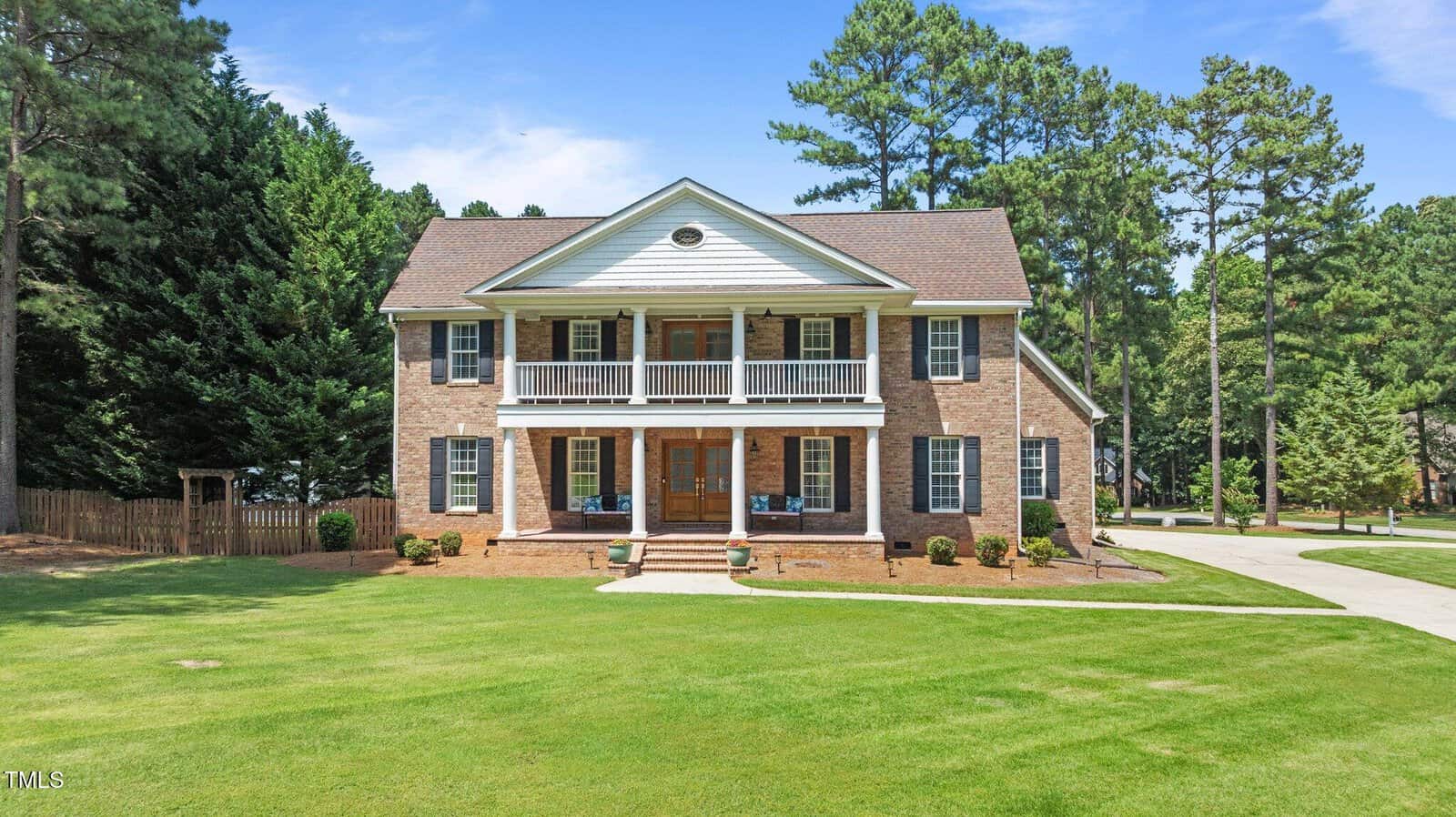 A two-story brick house with white trim and a double front porch, surrounded by a well-maintained lawn and tall trees, under a blue sky.