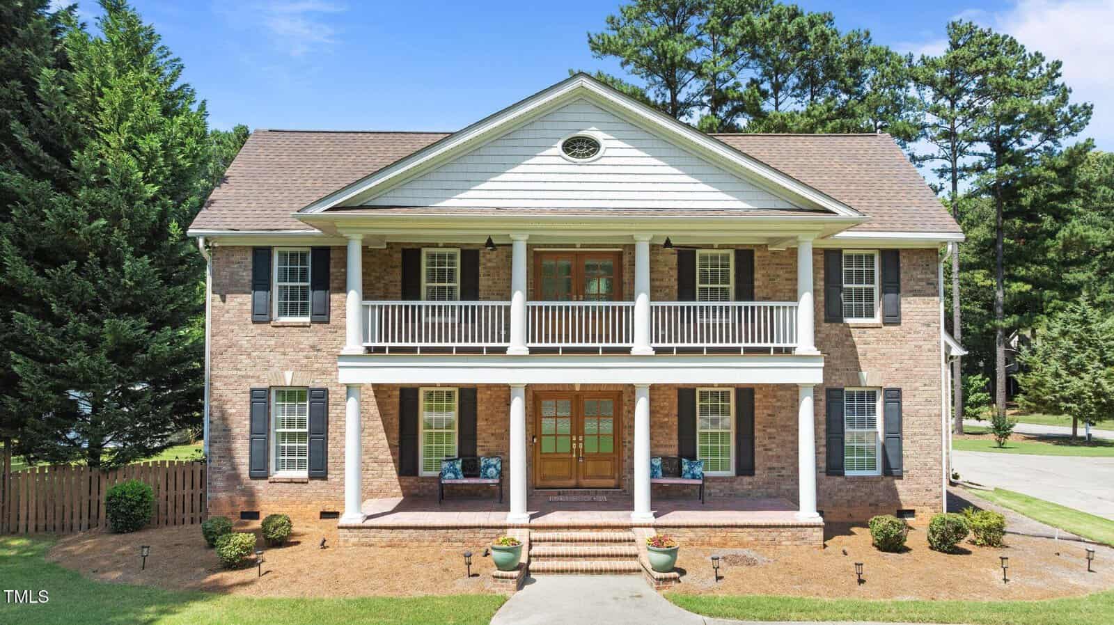 Two-story brick house with white columns, a covered front porch and balcony, wooden double front doors, and black shutters. The house is surrounded by green lawns, trees, and a driveway.