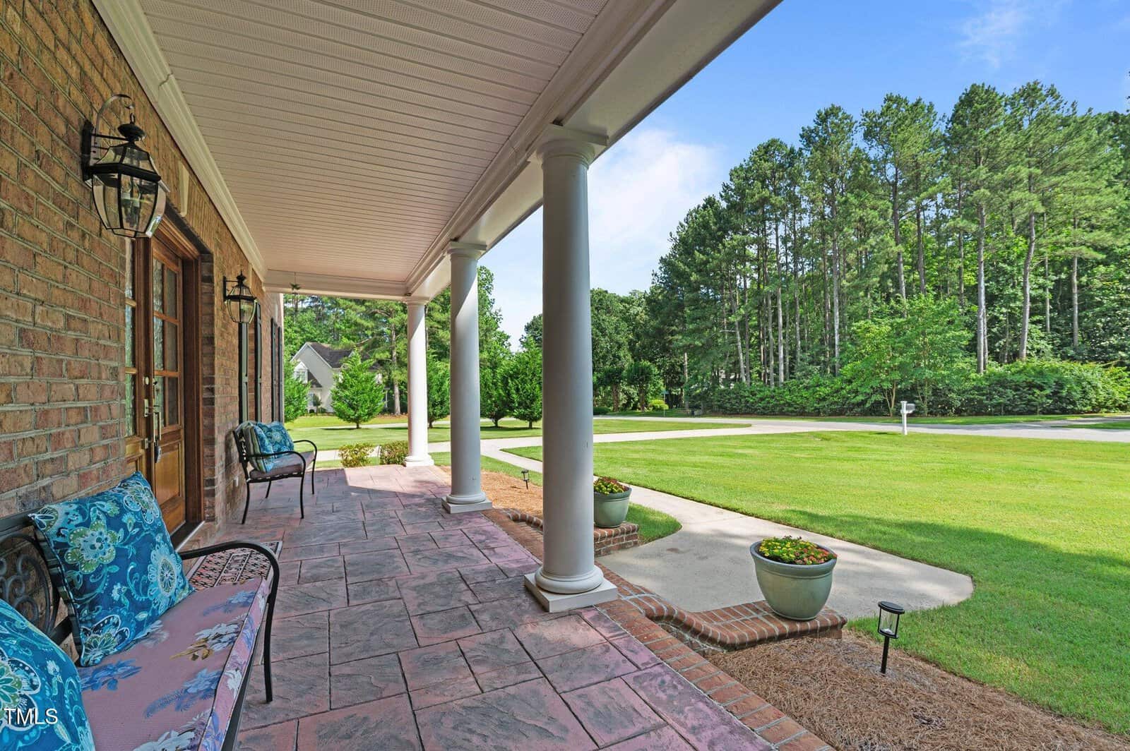 A spacious front porch with columns, patterned stone flooring, benches with blue cushions, potted plants, and a view of a green lawn, driveway, and tall trees under a blue sky.