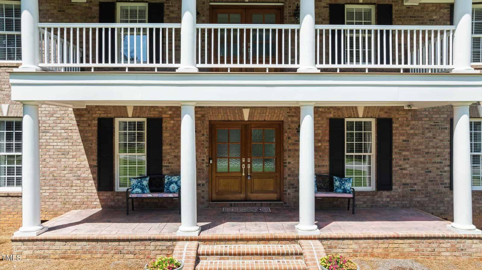 A brick house with a double wooden front door, white columns, a covered porch with two benches, and a second-story balcony featuring white railings. Steps lead up to the entrance with decorative plants on either side.