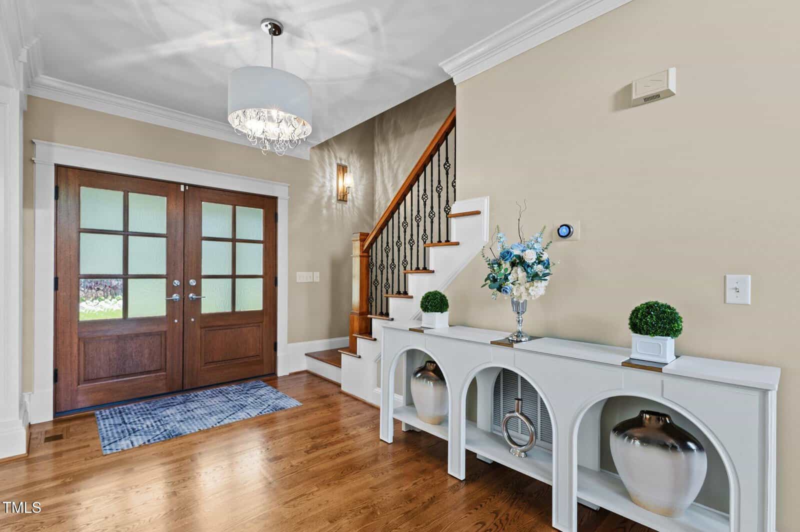 Entryway with double wooden doors, a staircase with iron balusters, a white console table holding two plants, two vases, and a floral arrangement. A chandelier hangs from the ceiling. The floor is hardwood, and there is a blue rug.