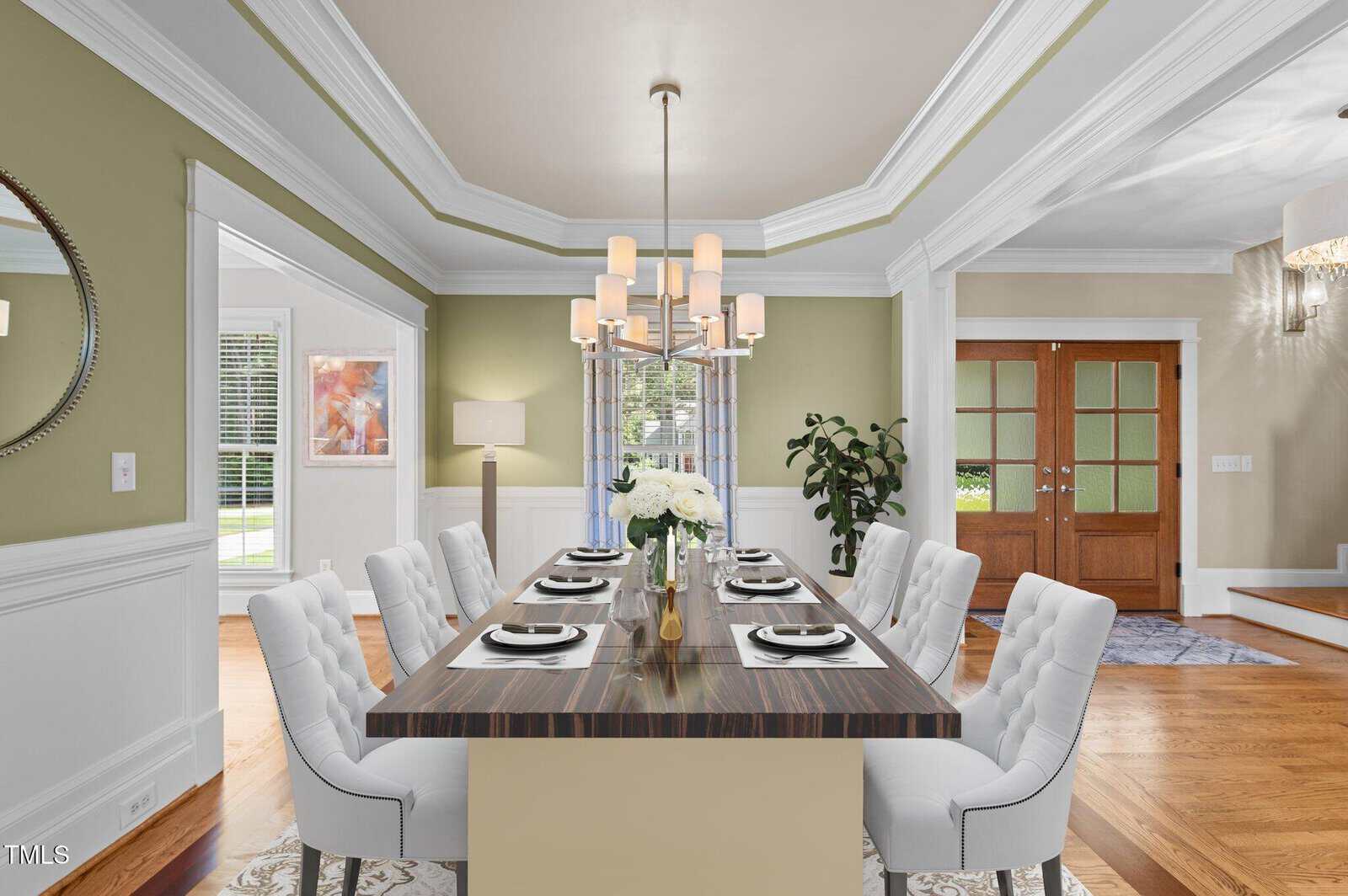 A dining room with a rectangular table set for eight, white cushioned chairs, a modern chandelier, wood flooring, green walls, and double glass doors. A large window and a potted plant are in the background.