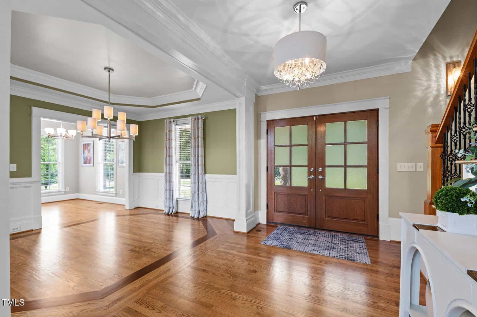 A spacious entryway with double wooden doors, a chandelier, hardwood floors, and an adjacent dining area with a modern light fixture and green accent wall. Large windows let in natural light.