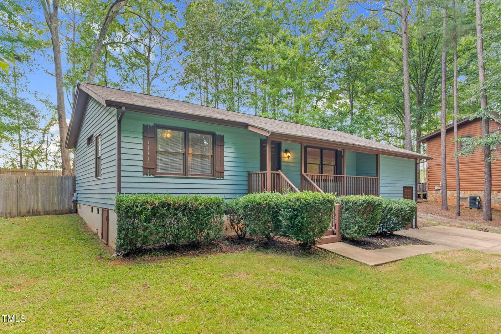 A single-story blue house with brown shutters and a covered front porch, surrounded by green bushes and trees. A concrete walkway leads to the steps. The lawn is neatly maintained and the house has a fenced backyard.