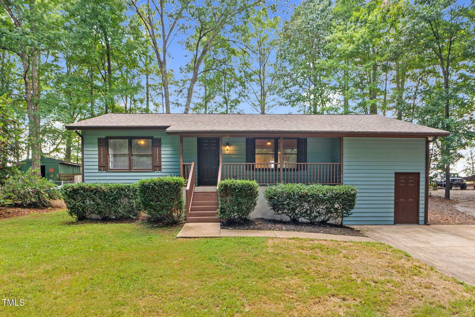This charming single-story light blue house at 424 Shawnee Drive, Louisburg, features brown shutters and a covered front porch. Shrubs line the front, and a concrete path leads to steps up to the porch. Trees and a grassy yard surround the house, creating a welcoming atmosphere.