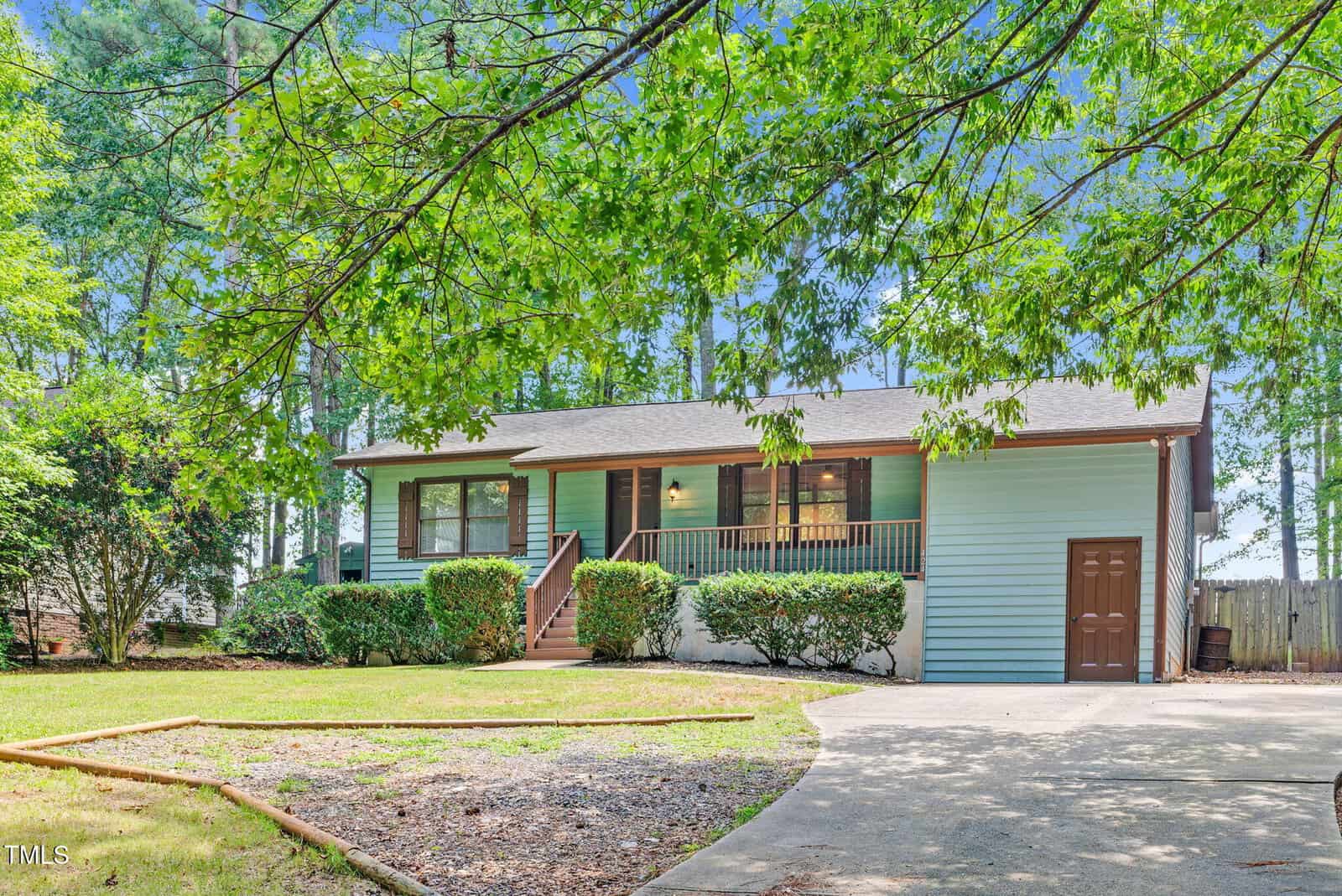A single-story house with light blue siding, a covered front porch, brown trim, and attached garage sits behind a curved driveway. Green shrubs line the front, and mature trees surround the yard.