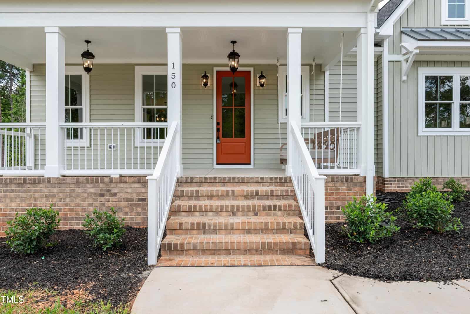 Front view of a house entrance with brick steps leading to a covered porch, white railings, three hanging lantern lights, green siding, a red front door, and small bushes along the walkway.