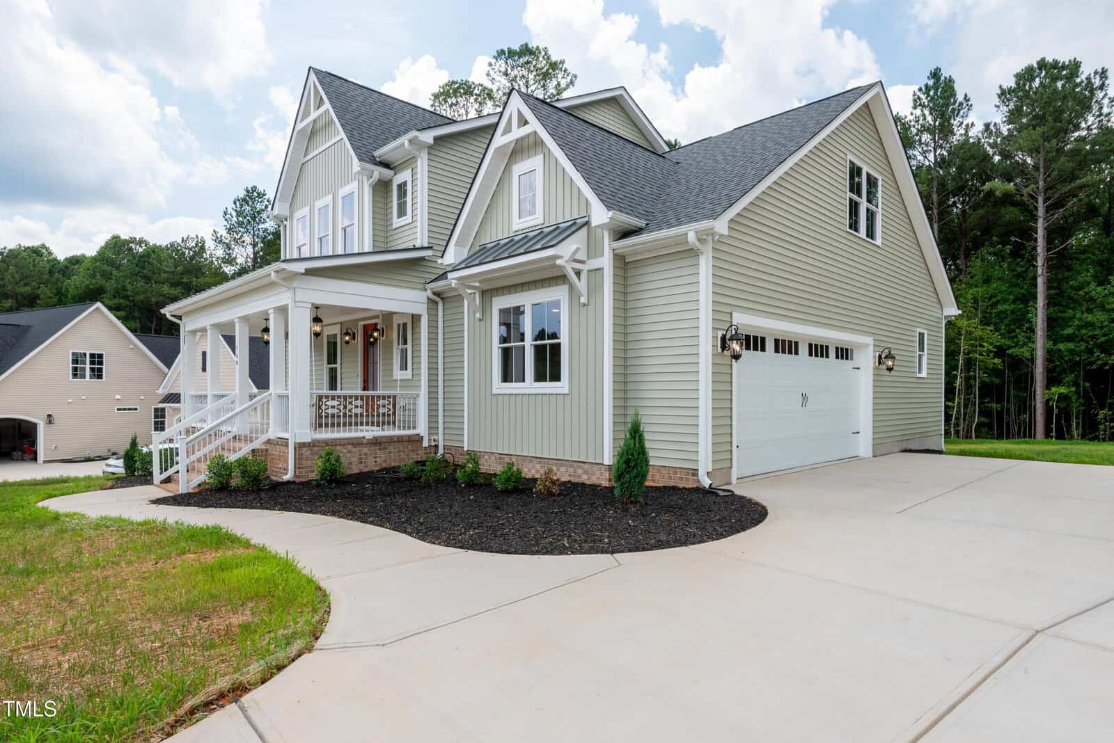 Two-story house with light gray siding, white trim, and a covered front porch. The house has a double garage, landscaped front yard with mulch and shrubs, and sits on a large concrete driveway. Tall trees are in the background.