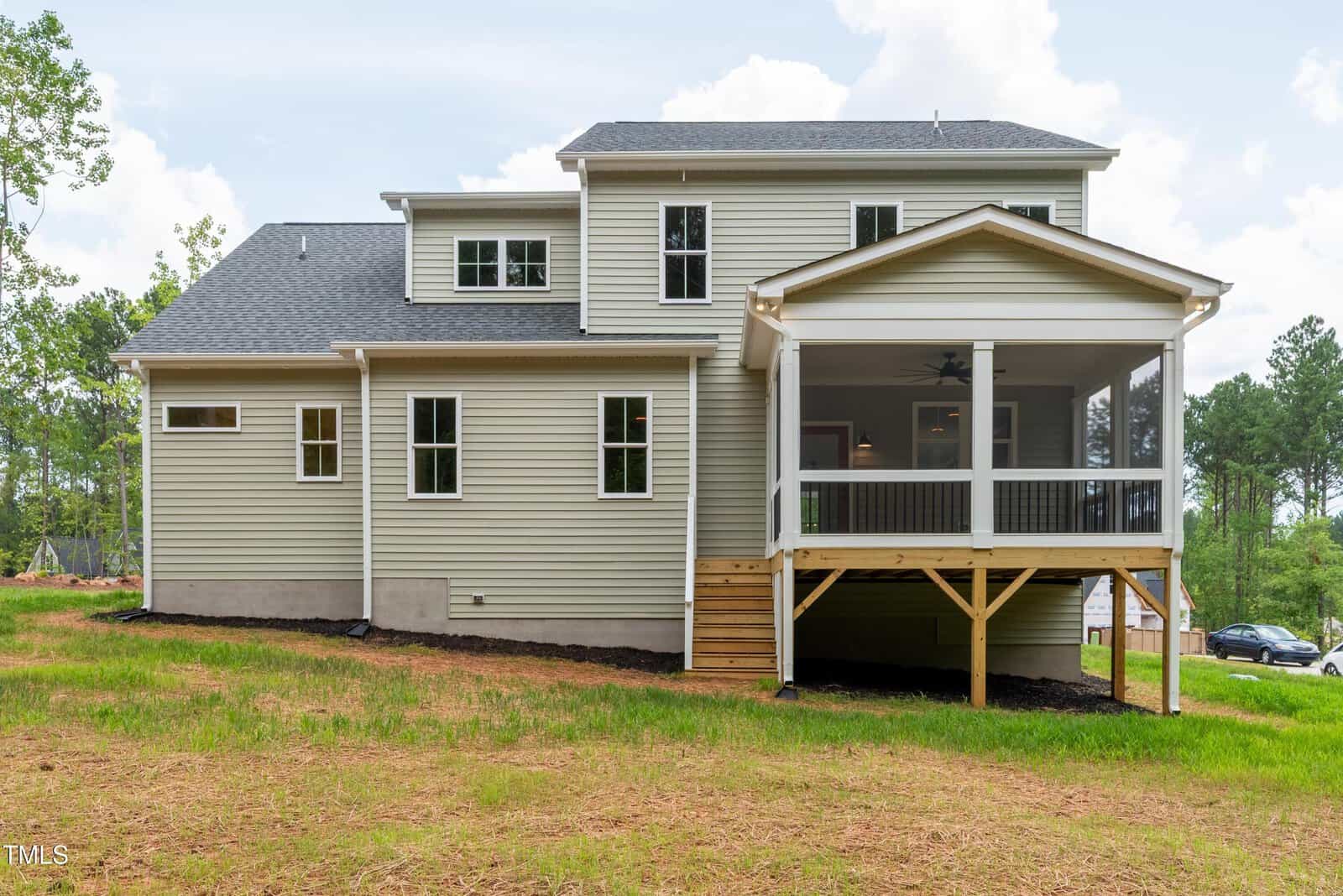 Two-story beige house with white trim, several windows, and a screened porch with a ceiling fan. The house is elevated with a set of wooden stairs leading to the porch. The yard has grass and is bordered by trees.
