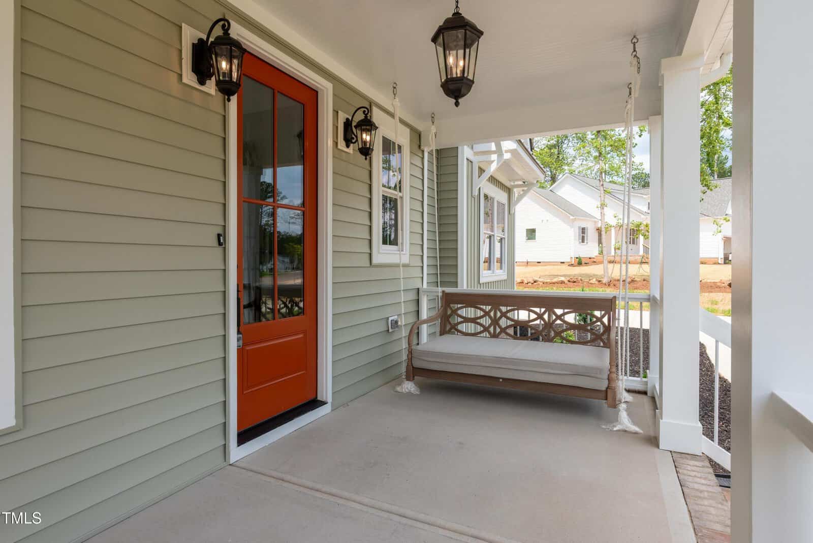 A covered front porch with light green siding, an orange front door with glass panes, black lantern-style lights, and a wooden porch swing with a cushion. Houses and landscaping are visible in the background.