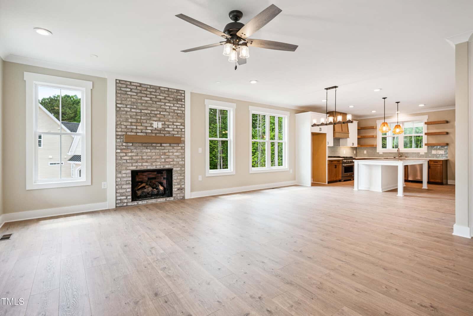 Spacious open-concept living area with light wood flooring, a ceiling fan, a stone fireplace, large windows, and a view of the kitchen with white counters and wood shelving.