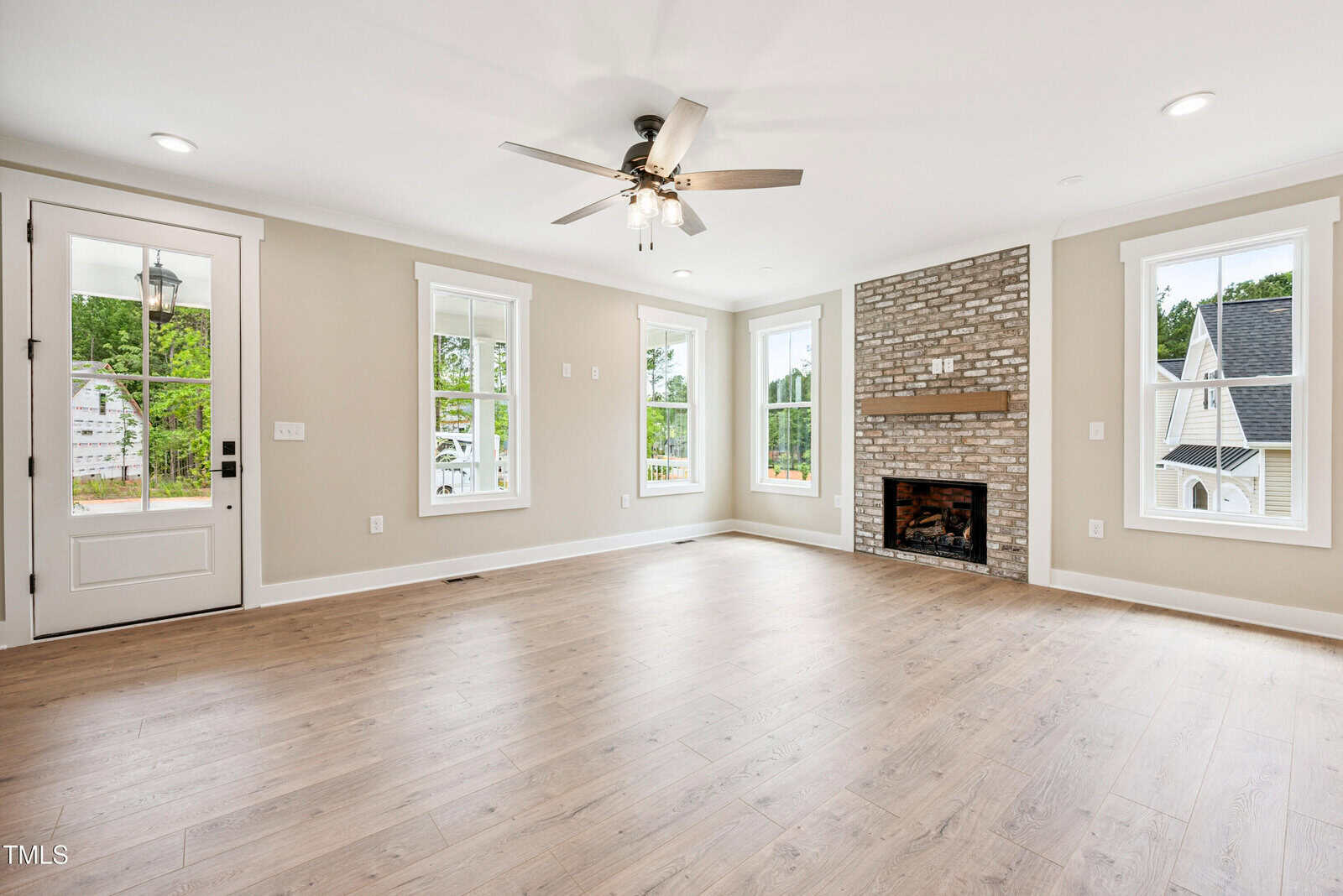 A bright, empty living room with light wood flooring, beige walls, multiple windows, a ceiling fan, a brick fireplace with a wood mantel, and a white front door with glass panels.