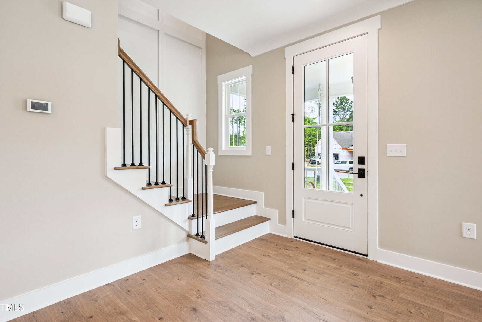 A modern entryway with light wood flooring, beige walls, a white front door with glass panels, a small window, and a staircase with wooden steps and black metal railings.