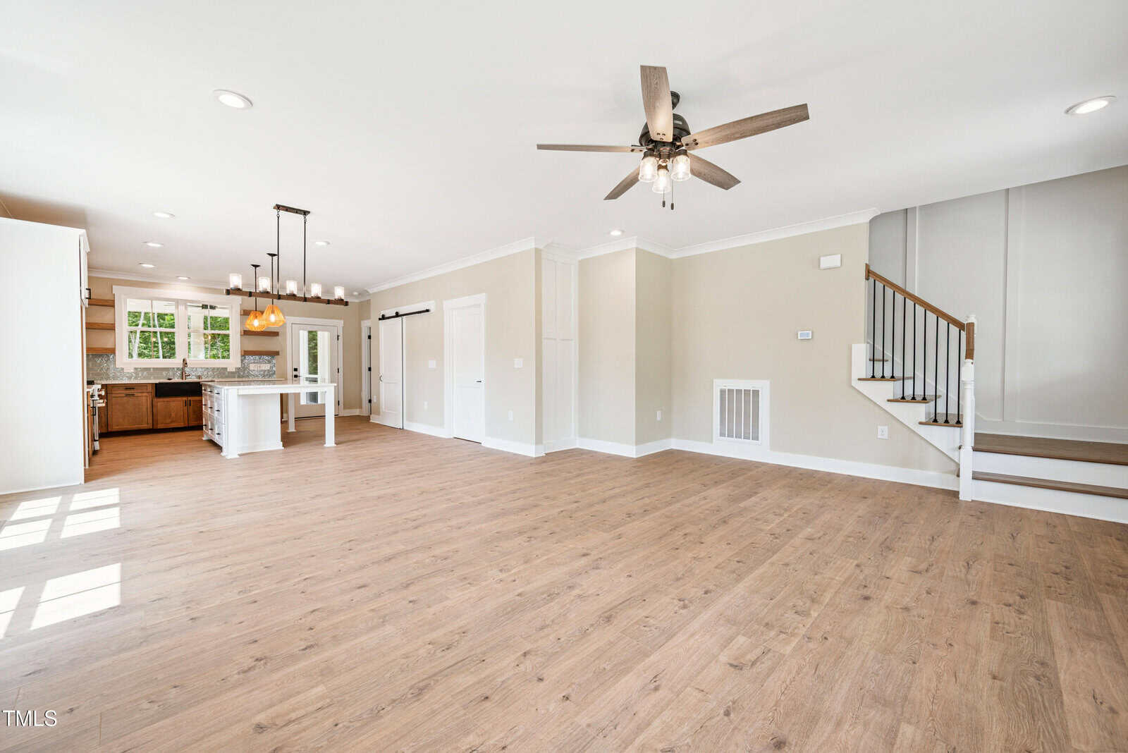 Open-concept living area with light wood flooring, neutral walls, a ceiling fan, recessed lighting, a modern kitchen with white cabinets and island, and a staircase with a dark handrail and white balusters.