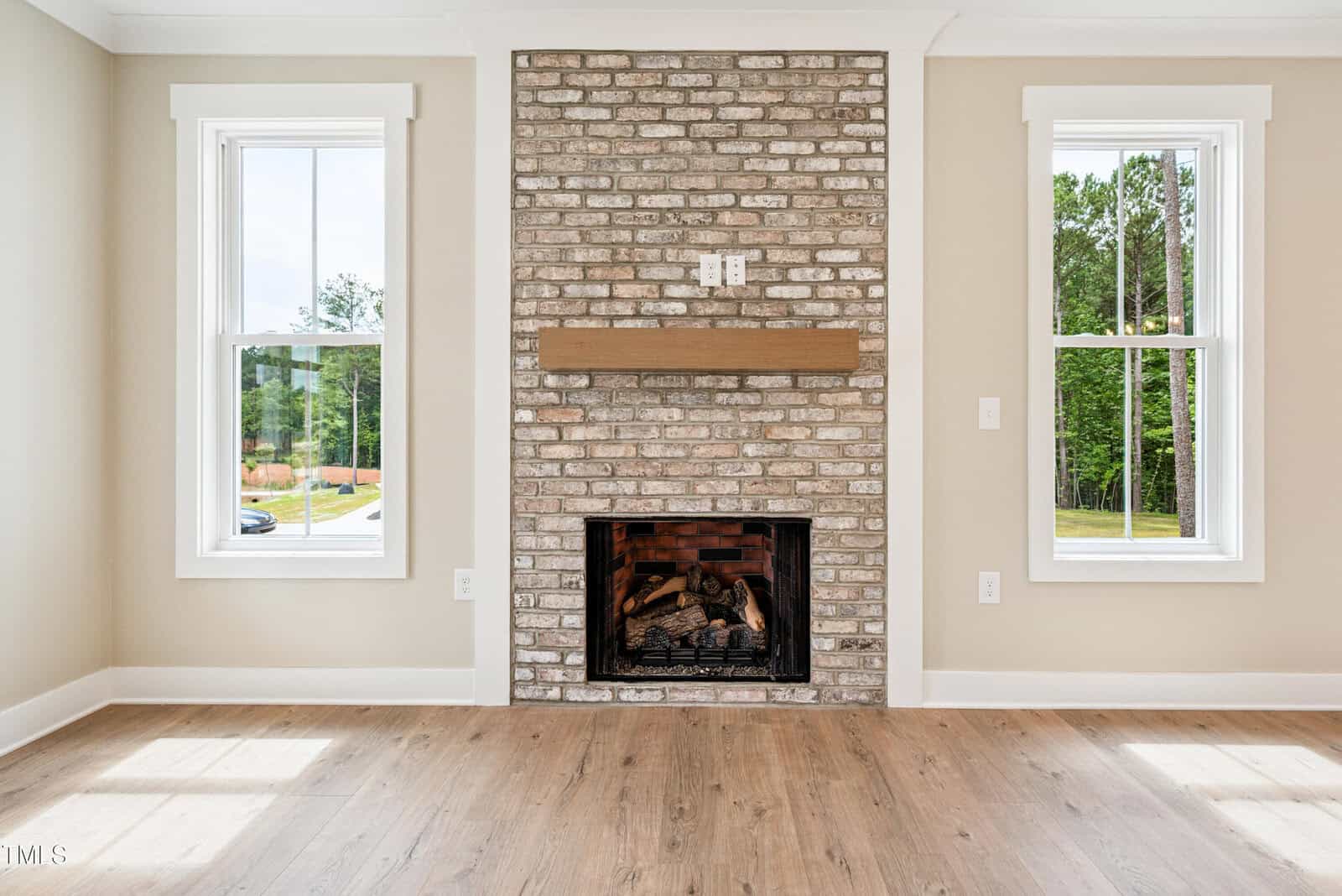 A living room with a brick fireplace and wooden mantel is centered between two tall windows. The room has light-colored walls and wood floors, with greenery visible outside.
