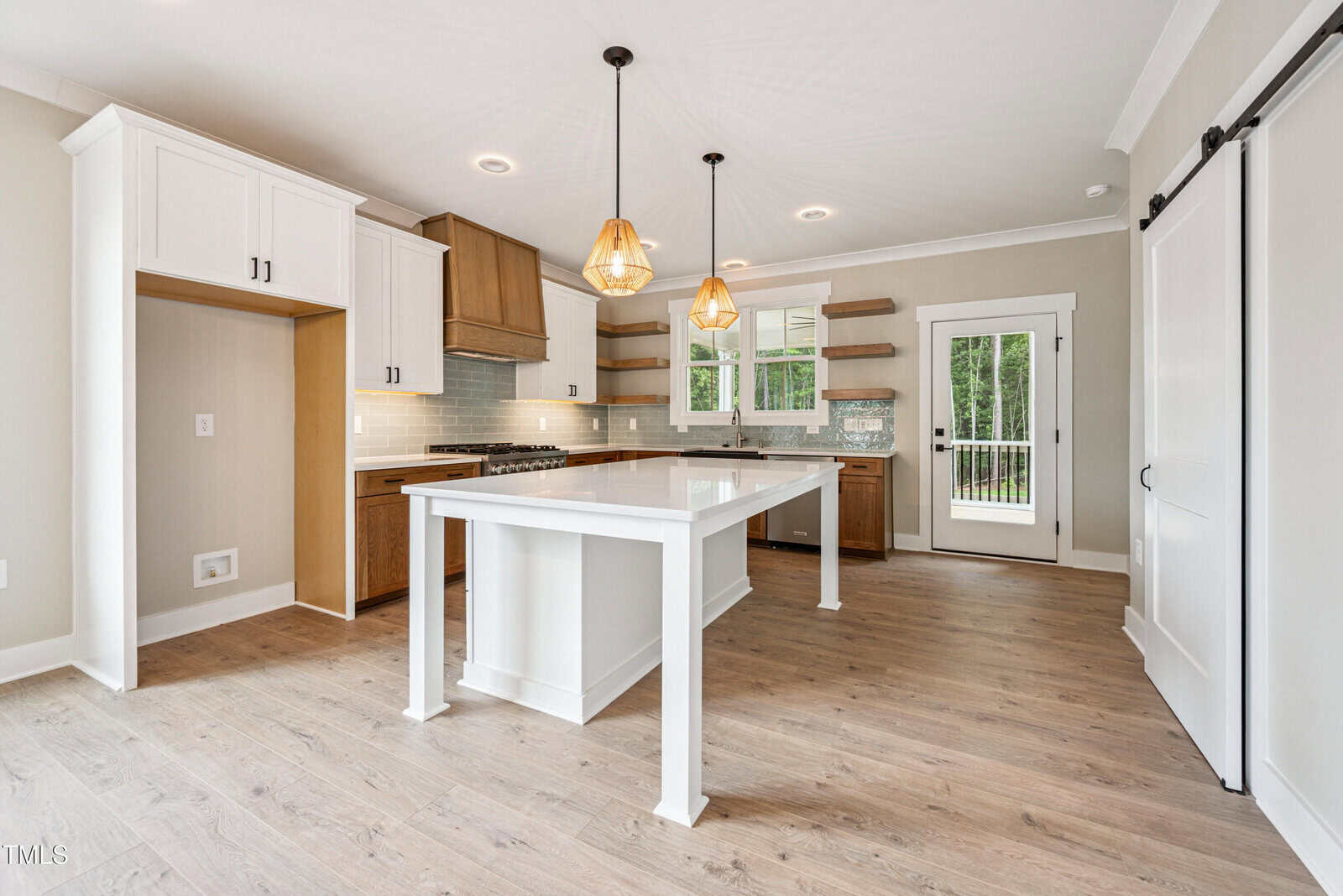 Modern kitchen with wood floors, white cabinets, a large center island, glass pendant lights, open shelves, stainless steel appliances, and a sliding barn door leading to a bright room with windows and a door to the outside.