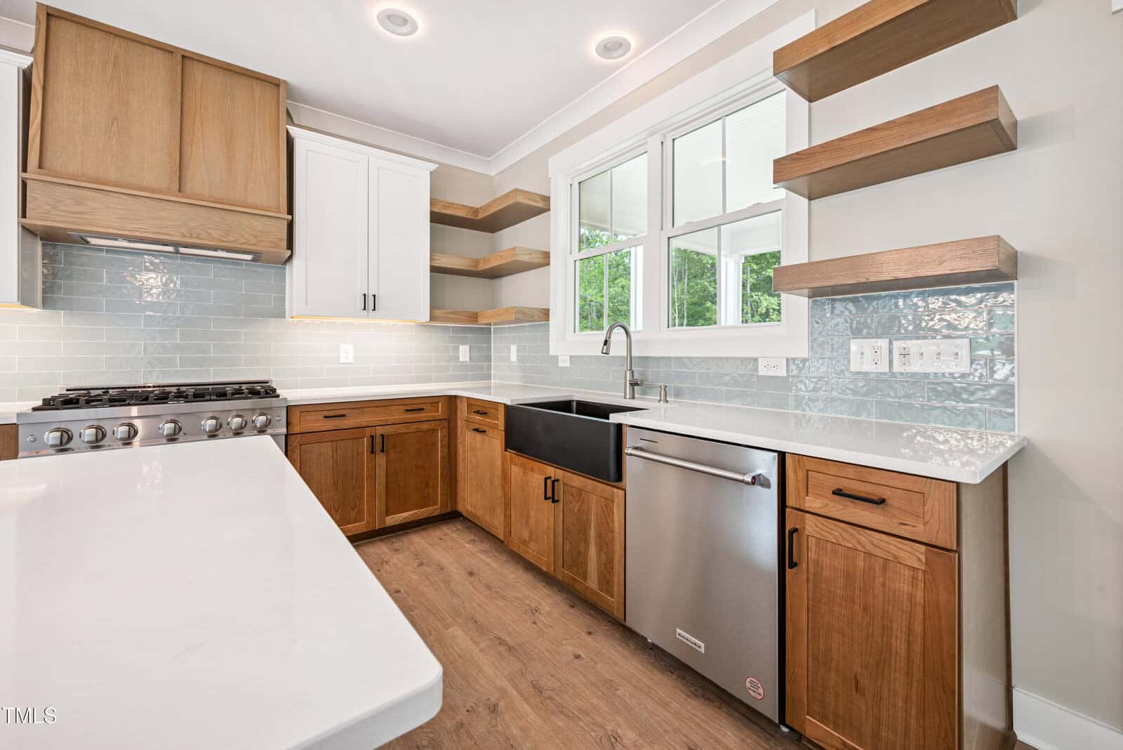 Modern kitchen with wood cabinets, white countertops, open wood shelving, a stainless steel dishwasher, farmhouse sink, gas stove, light blue subway tile backsplash, and large window above the sink.