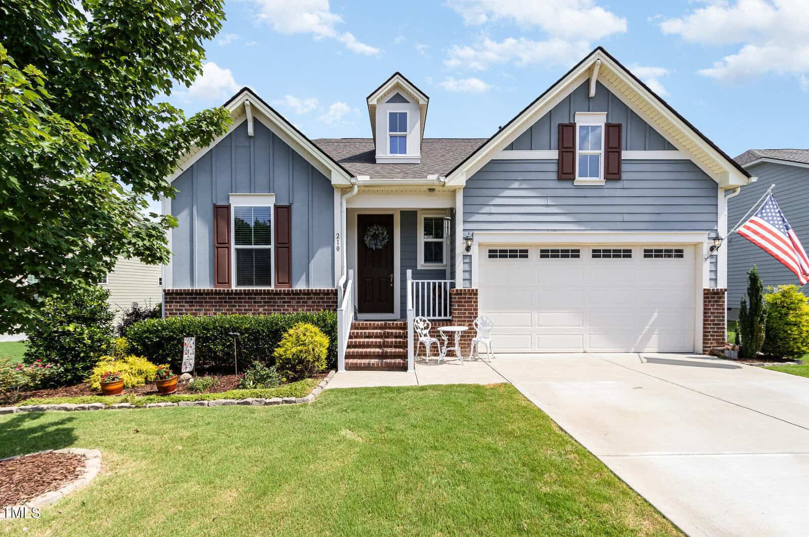 A gray single-story house with white trim, brick accents, a two-car garage, red shutters, a small front porch with chairs, a wreath on the door, an American flag, and a well-maintained lawn and garden awaits you at 210 Olde Liberty Drive in Youngsville.