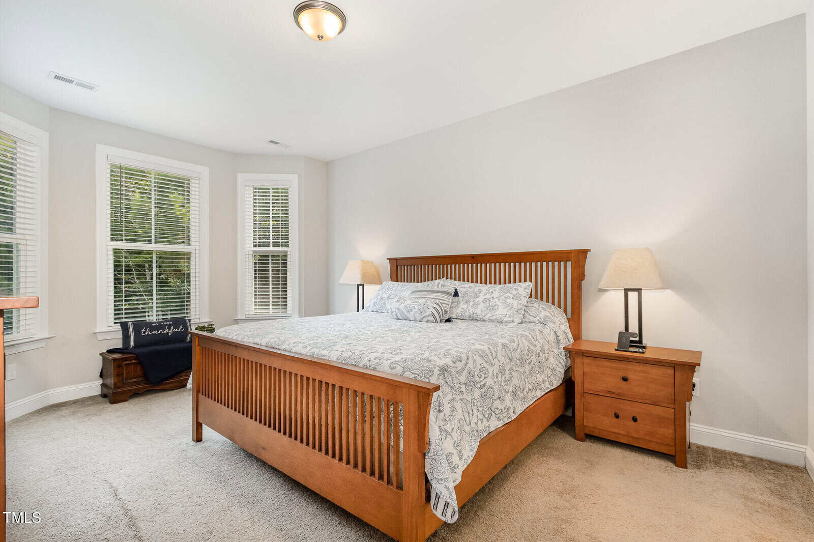 A bedroom with a wooden bed frame, matching nightstands, two lamps, light walls, beige carpet, and three large windows. A dark chair and small table are in the corner near the windows.