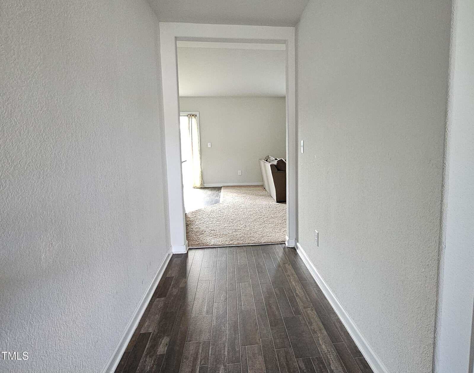 A hallway with dark wood flooring leads to a carpeted living room area. A beige armchair is visible near a sliding glass door with light coming in. The walls are white and minimalistic.