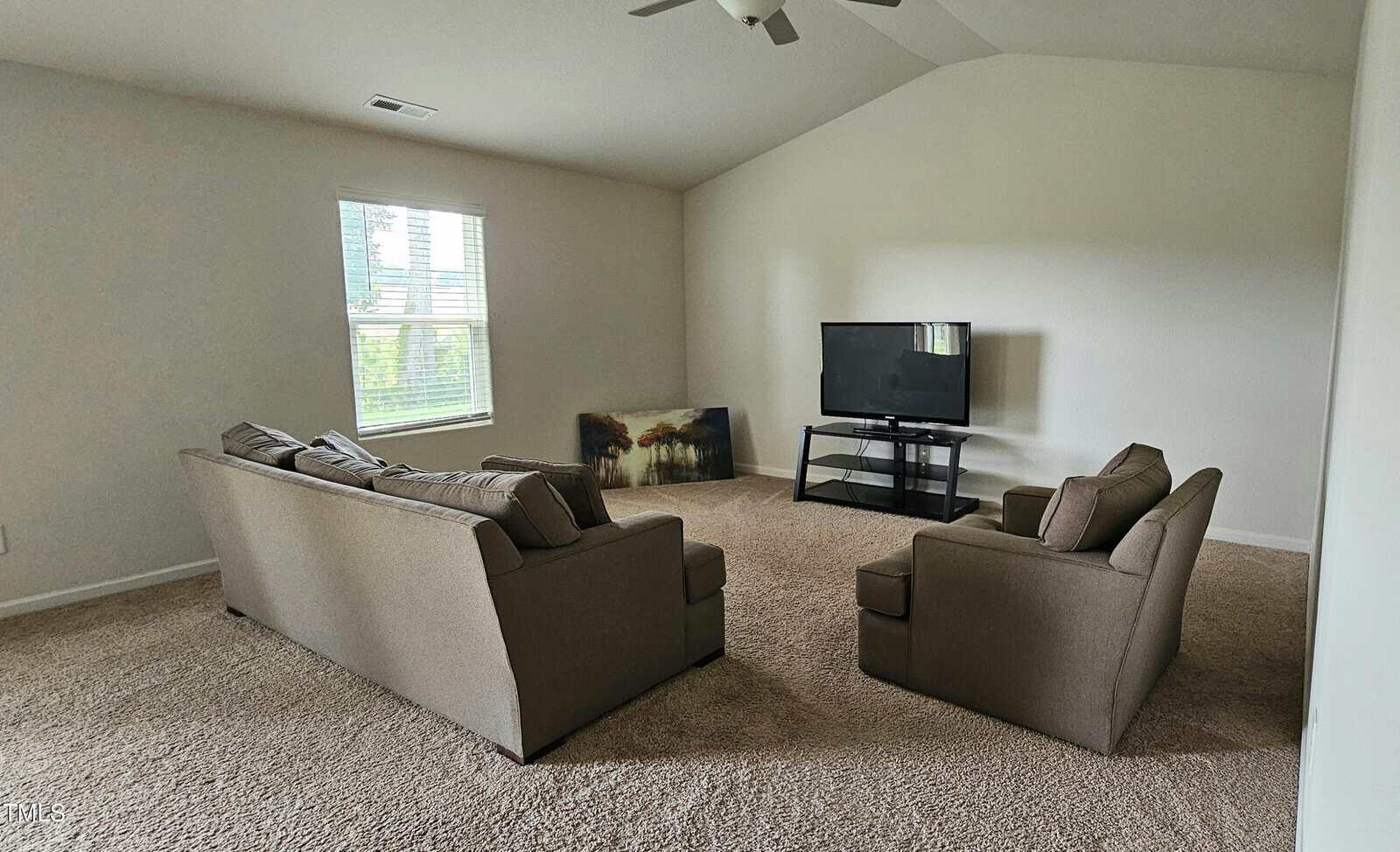A living room with beige carpet, a sofa, and an armchair facing a TV on a stand. A painting rests against the wall. There is one window with light coming in. The walls are plain and light-colored.