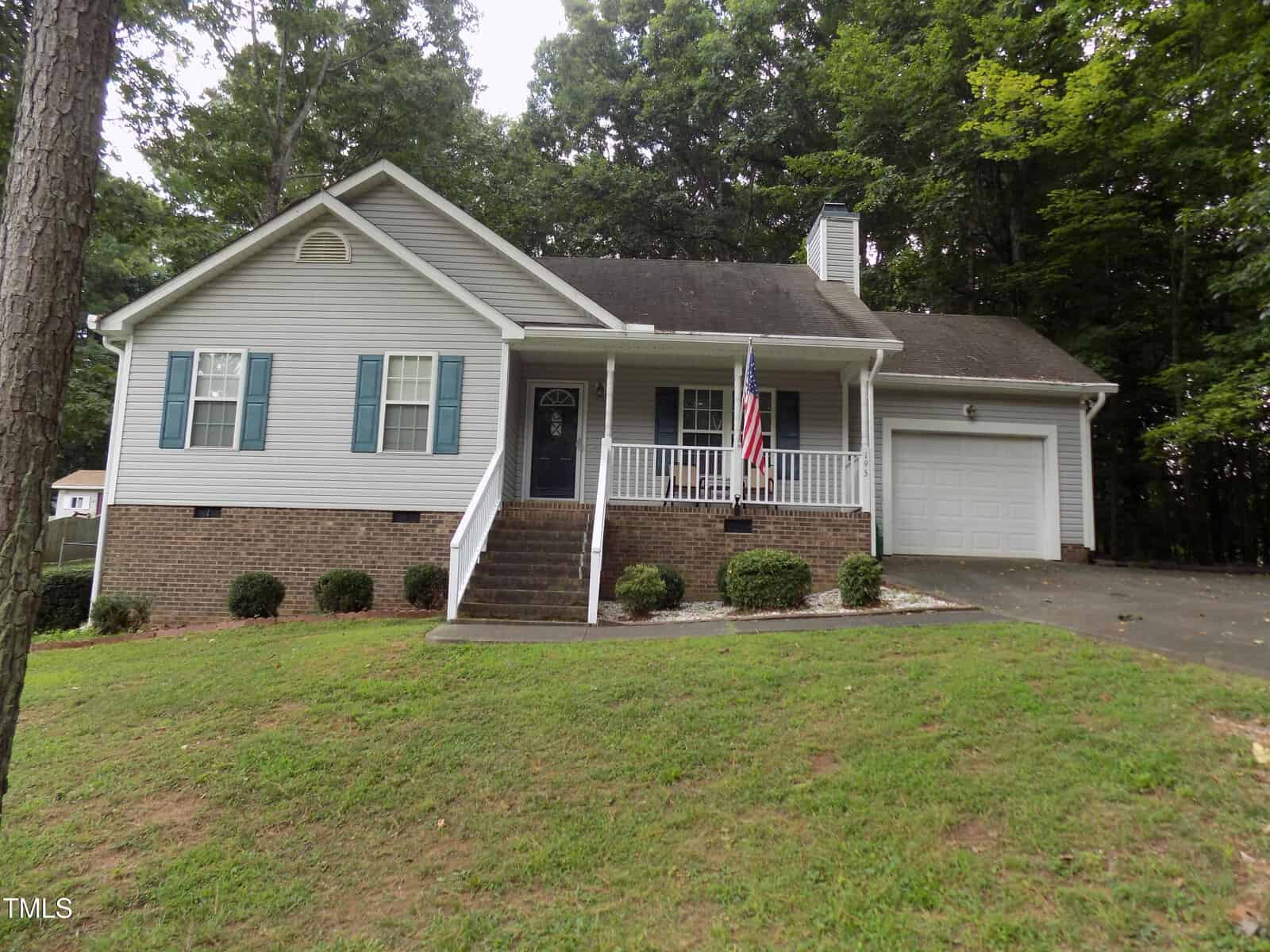 Single-story house with gray siding, blue shutters, and a brick foundation. There is a front porch with white railings, a U.S. flag, front steps, a driveway, garage, and a lawn with shrubs. Trees surround the house.