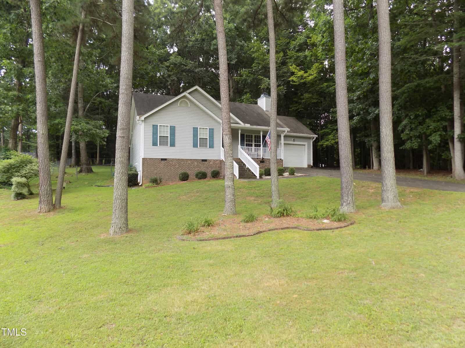 Single-story house with light gray siding, brick foundation, white shutters, and a front porch with steps. Nestled on 195 Eagle Stone Ridge in Youngsville, the home sits on a grassy lawn surrounded by tall trees and is bordered by a wooded area.