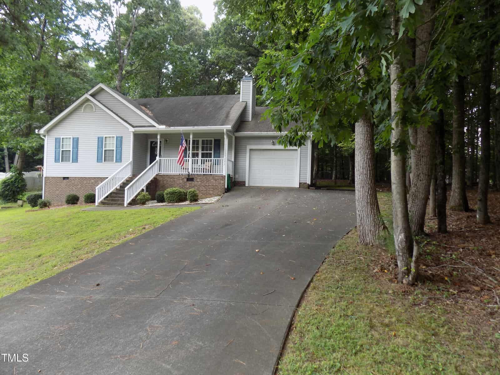 A single-story house with a front porch, attached garage, and American flag. The driveway is sloped and surrounded by grass and trees. The house is light-colored with brick accents and white railings.