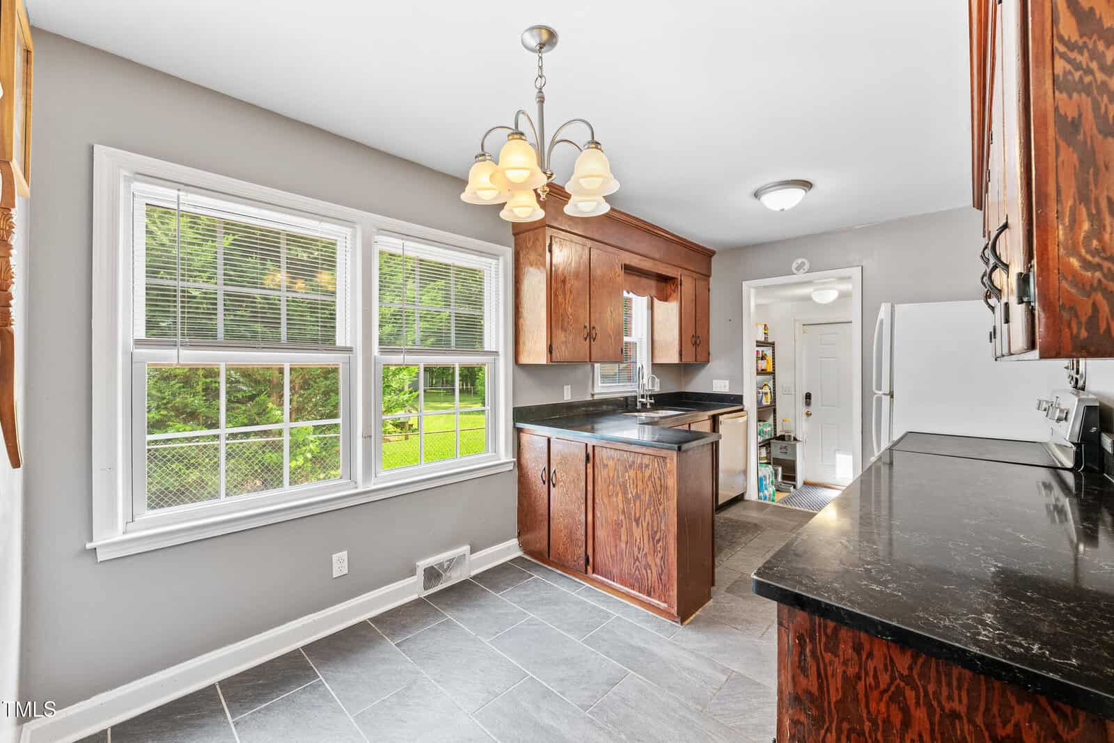 Kitchen with dark wood cabinets, black countertops, gray tile floor, and large window overlooking a grassy backyard. Refrigerator and door to adjacent room visible in the background. Ceiling light fixture above.