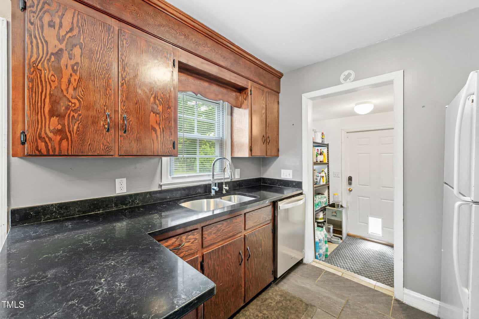 Kitchen with dark wood cabinets, black countertops, stainless steel sink and dishwasher, window above the sink, light gray walls, tile floor, and a view into a pantry and laundry area near a back door.