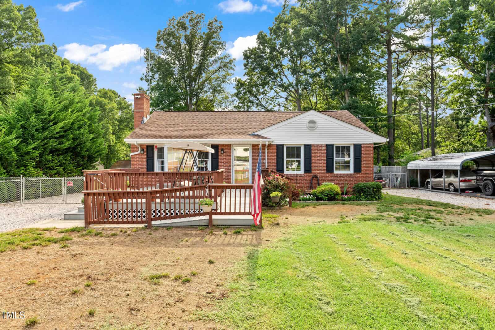 A single-story brick house with a front porch, American flag, and ramp, set in a green yard with trees and a gravel driveway. There are two vehicles parked under a carport to the right.