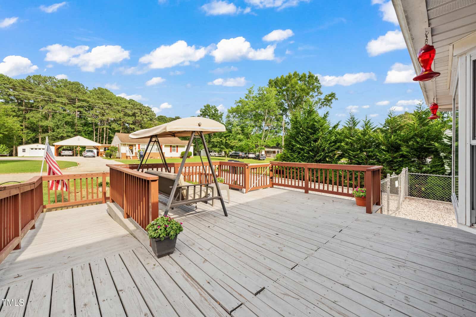 A spacious wooden deck with a porch swing, potted plants, and red hummingbird feeders overlooks a green yard with trees, a flag, and several detached buildings under a blue sky with scattered clouds.